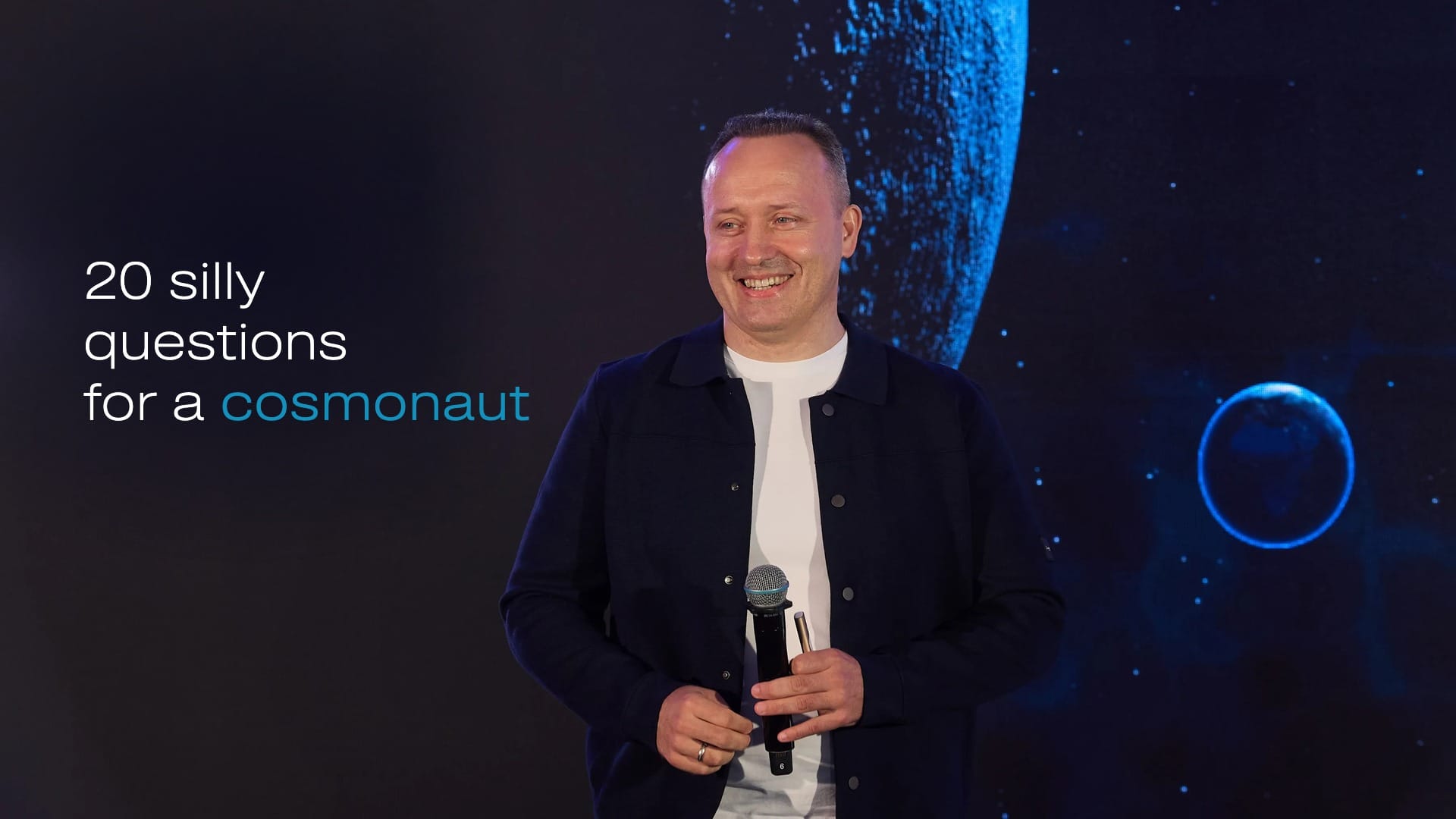 Cosmonaut Alexander Misurkin on stage with a microphone; caption «20 silly questions for a cosmonaut»